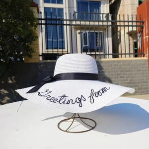 White wide brim straw hat with Greetings from embroidery and black ribbon bow detail
