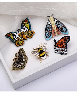 Detail showing five insect enamel pins including moths and bees on white fabric background