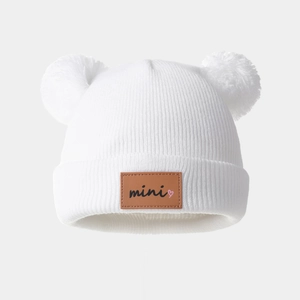 Detailed knit texture of white baby hat featuring fluffy double pom poms and mini patch