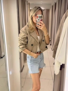 Detail view of a beige double-breasted cropped trench coat styled with denim shorts