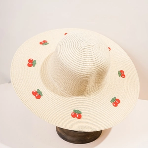 Dettaglio ravvicinato dei ricami a ciliegia rossa su un cappello estivo in paglia beige
