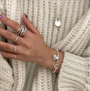 Detail of silver heart bracelet and layered vintage rings on a beige sweater background