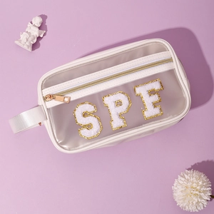 Dettaglio borsa cosmetica in PVC con toppe SPF in ciniglia dorata e rifiniture bianche