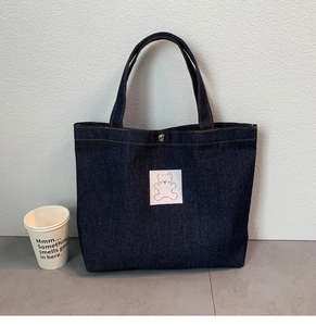 Detail of dark blue denim tote bag with white bear patch and side size comparison cup