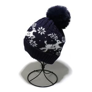 Detail of dark blue knit beanie featuring white reindeer patterns and a fluffy pom-pom
