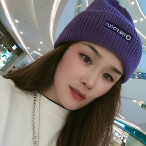 Close-up of a deep purple knitted beanie with a THE FOCUS patch in a shopping mall setting