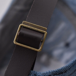 Detail view of the brown leather adjustment strap and bronze buckle on a denim baseball cap