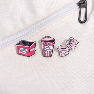 Vue rapprochée de trois épingles en émail rose avec motifs de toilettes et poubelles
