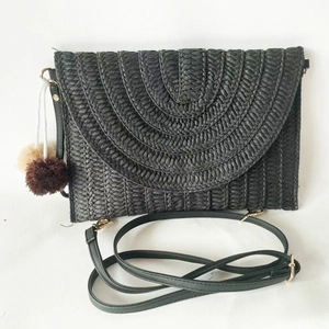 Detail of black woven envelope straw bag featuring bicolor pompom charm and shoulder strap