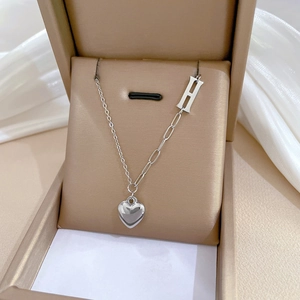 Close-up of silver steel necklace featuring an H-letter charm and heart pendant in a box
