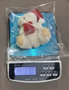 Close-up of small white plush Christmas bear with red hat holding a gift box in packaging