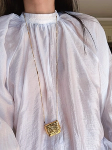 Detailed close-up of a long golden necklace featuring a rectangular woven texture pendant