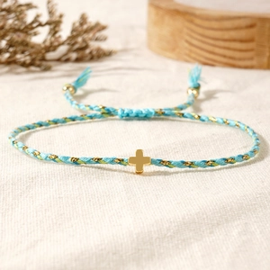 Detail of hand-woven light blue cord bracelet with a gold cross charm on linen fabric