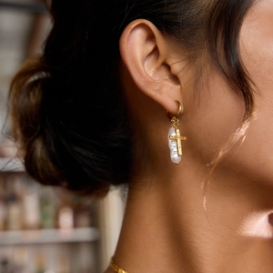 Close-up of golden cross and baroque pearl earrings worn by a model in warm light