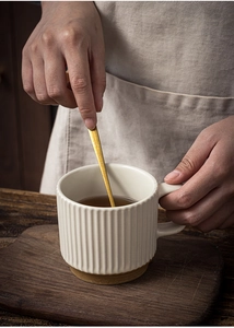 Close-up of hands stirring coffee in a cream ceramic mug with wood coaster and gold spoon
