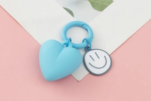 Detailed view of a blue 3D heart keychain with a grey smiley face charm on pink background