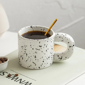 Minimalist splatter ink ceramic mug with golden spoon and coffee beans on white table