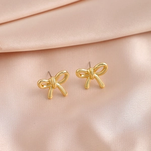 Close-up of golden metal bow earrings displayed on a soft pink silk fabric background