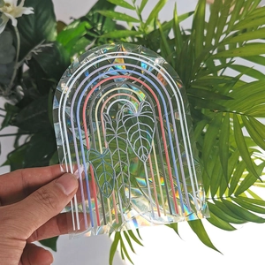 Hand holding holographic suncatcher sticker with rainbow arch and leaf design on window