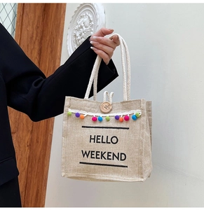Close-up of woven jute tote bag with HELLO WEEKEND text and colorful pompom fringe