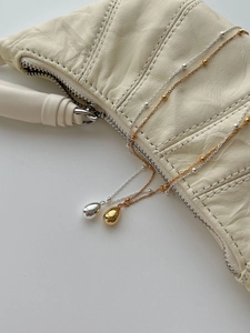 Detail of minimalist gold and silver teardrop pendants on bead chains over a cream leather bag