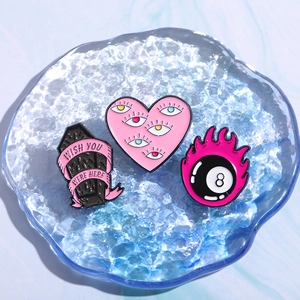 Three trendy enamel pins with pink heart, coffin, and 8-ball designs in a blue glass tray
