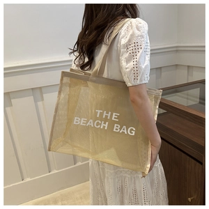 Model showing a beige mesh beach tote bag with THE BEACH BAG text in a minimalist setting