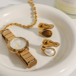 Dettaglio di anelli dorati con perla e occhio di tigre accanto a un orologio di moda