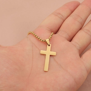 Polished gold stainless steel cross pendant necklace held in hand over pink fabric