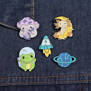 Close-up of five cute enamel pins featuring a frog, mushroom, and space on denim fabric