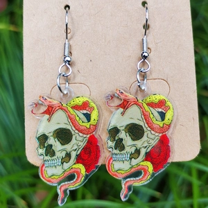 Nahaufnahme der Acryl-Ohrringe mit Totenkopf-, Schlangen- und Rosenmotiven auf Gras