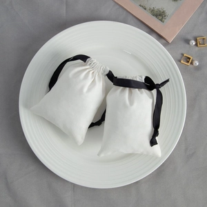 Pair of white cotton drawstring jewelry bags with black silk ribbons on a white ceramic plate