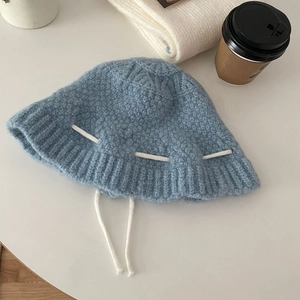 Flat lay of a blue knitted bucket hat with white bow ribbons on a minimal white desk
