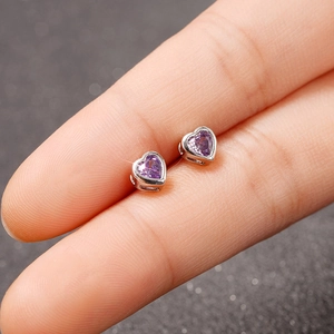 Close-up of a silver heart stud earring with a purple gemstone held by a finger