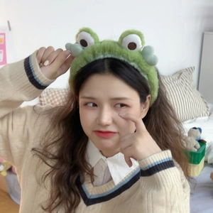 Close-up of a plush green frog headband with large eyes worn by a model in a cozy setting