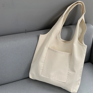 Blank beige canvas tote bag with front pocket leaning on a soft gray fabric sofa