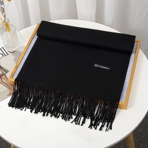 Detail of a black cashmere-feel scarf with tassels and logo tag on a minimalist table