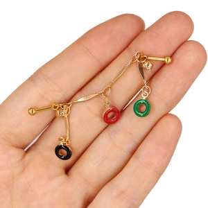Gold barbell piercing with red, green, and black drop charms shown in hand for scale