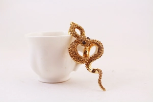 Detail of a golden snake keychain with brown rhinestones perched on a white ceramic cup