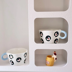 Close-up of ceramic penguin mug with irregular rim and cat ornament on a white shelf