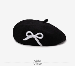 Detail of a black knit beret featuring white bow embroidery and ribbed vertical texture