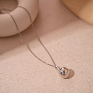 Pendentif sphérique en argent poli sur une chaîne fine posé sur une surface beige texturée