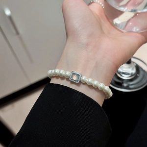 Elegant pearl bracelet with gold-plated square pendant shown on wrist while holding a cup