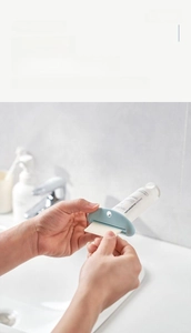 Detail showing hands operating a light blue manual toothpaste squeezer in a bathroom