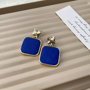 Detail of geometric gold earrings featuring blue velvet square charms on a textured tray