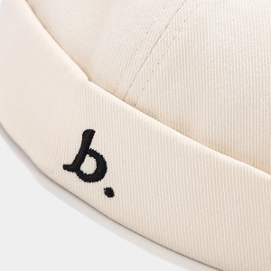 Gedetailleerde close-up van het zwart geborduurde 'b.' logo op een beige katoenen docker muts