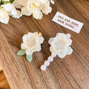 Close-up of two white floral hair clips with green leaves and pearl beads on wooden table