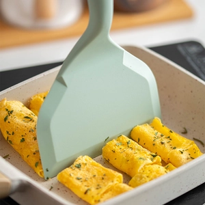 Silicone tamagoyaki spatula with wooden handle in mint green on a beige square frying pan