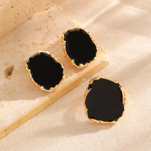 Detail of irregular black enamel earrings with gold-tone edges on a beige stone surface