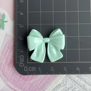 Macro detail of a 3.5cm light green resin bow charm next to a ruler for size scale